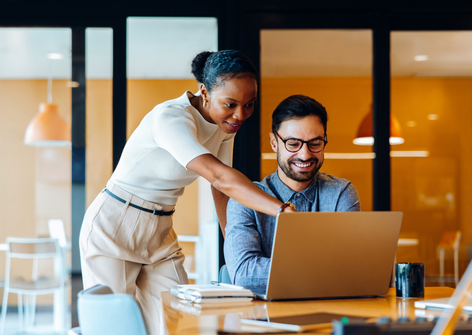 Two young professionals, a man and a woman, share ideas while reviewing work on a laptop in a corporate office setting, showcasing collaboration, productivity, and engagement at work.